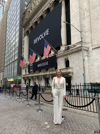 Woman in a white blazer and trousers standing in front of the New York Stock Exchange, with a large REVOLVE banner displayed on the building facade.
