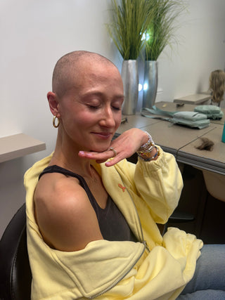 Young woman with a shaved head during treatment, eyes closed in a quiet smile, wearing a yellow hoodie with a cancer awareness ribbon pin and stacked bracelets.