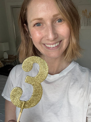 POST SWIM founder Lauren Yerkes smiles holding a gold glitter number 3, celebrating Triple Negative Breast Cancer Day.