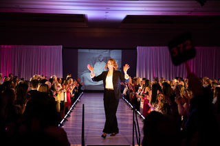 Lauren Yerkes walking the runway at the Runway for Recovery fashion show, arms raised to an applauding crowd, wearing a black blazer and trousers under pink-lit ballroom lighting.