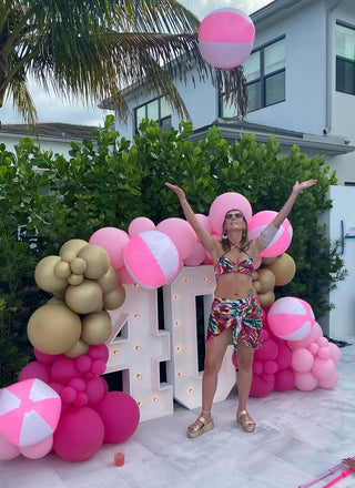 Nicholle celebrating her 40th birthday with arms raised, wearing a tropical print bikini and sarong, surrounded by a pink and gold balloon arch with illuminated "40" marquee letters.