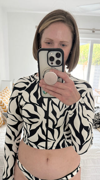 Woman taking a mirror selfie wearing a black and white printed POST SWIM long-sleeve zip-front rash guard crop top, showcasing the scar-coverage design.