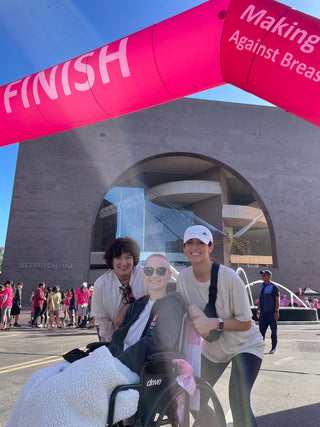 Three women smiling at the finish line of a breast cancer walk outside Segerstrom Hall, one seated in a wheelchair, united in support.