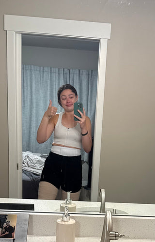 Woman taking a mirror selfie giving a thumbs up, wearing a post-surgical compression bra and binder, with tears in her eyes — a candid moment of resilience after breast surgery.