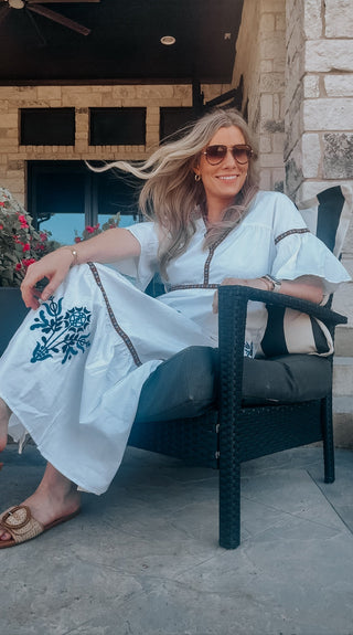 Woman with long blonde hair and aviator sunglasses relaxing on a patio chair, wearing a white embroidered dress with floral detail, smiling in the breeze.